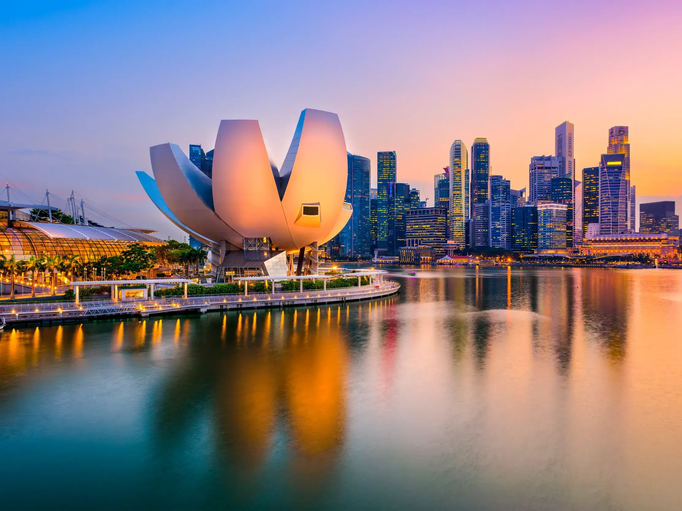 Das lotusförmige ArtScience Museum vor der Skyline Singapurs bei Dämmerung, die sich im Wasser der Marina Bay spiegelt.