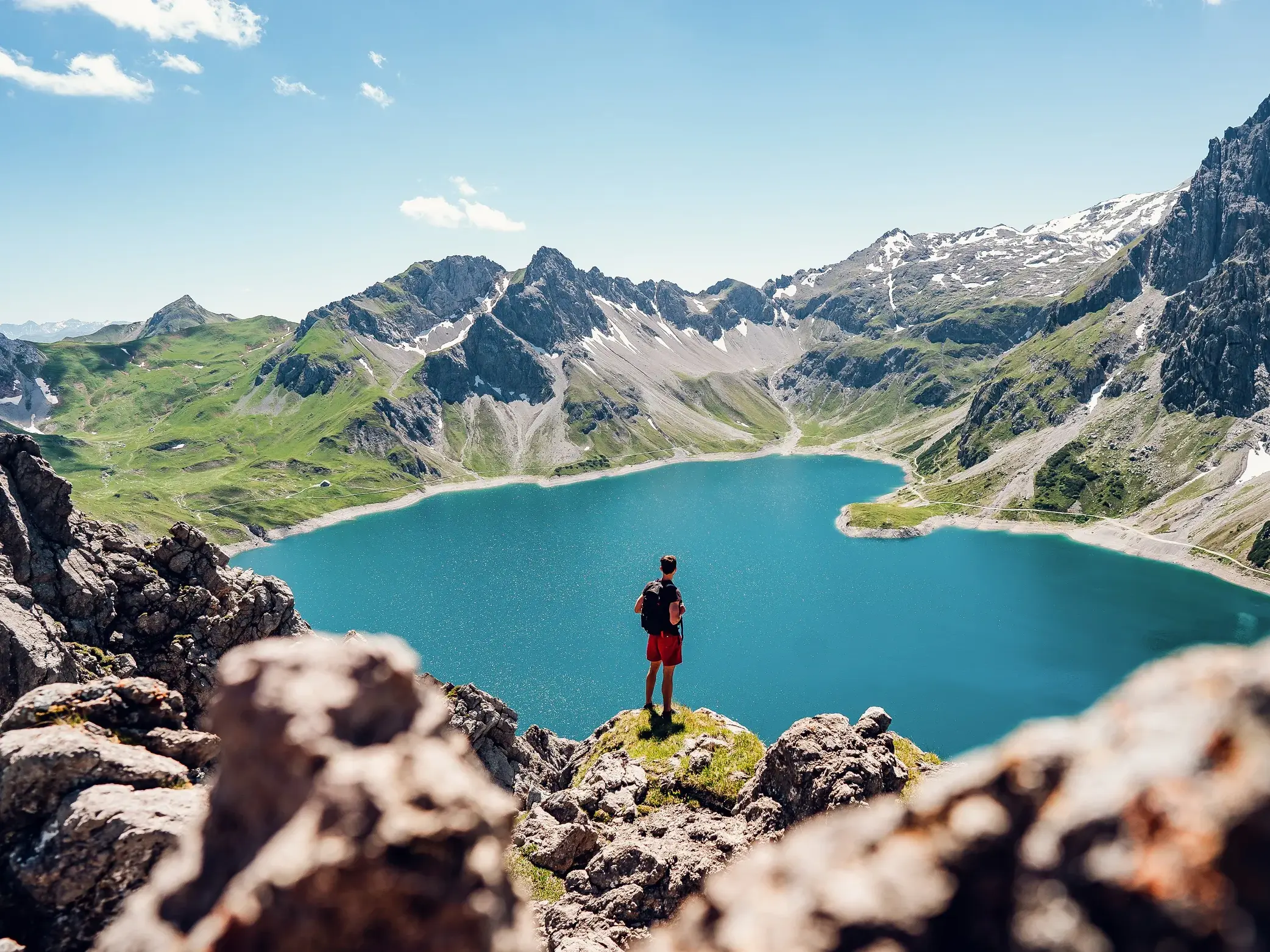 Ein Wanderer mit Rucksack genießt auf einem Felsen den Ausblick auf einen türkisen Bergsee und die Gipfel des Maira-Tals, Piemont.