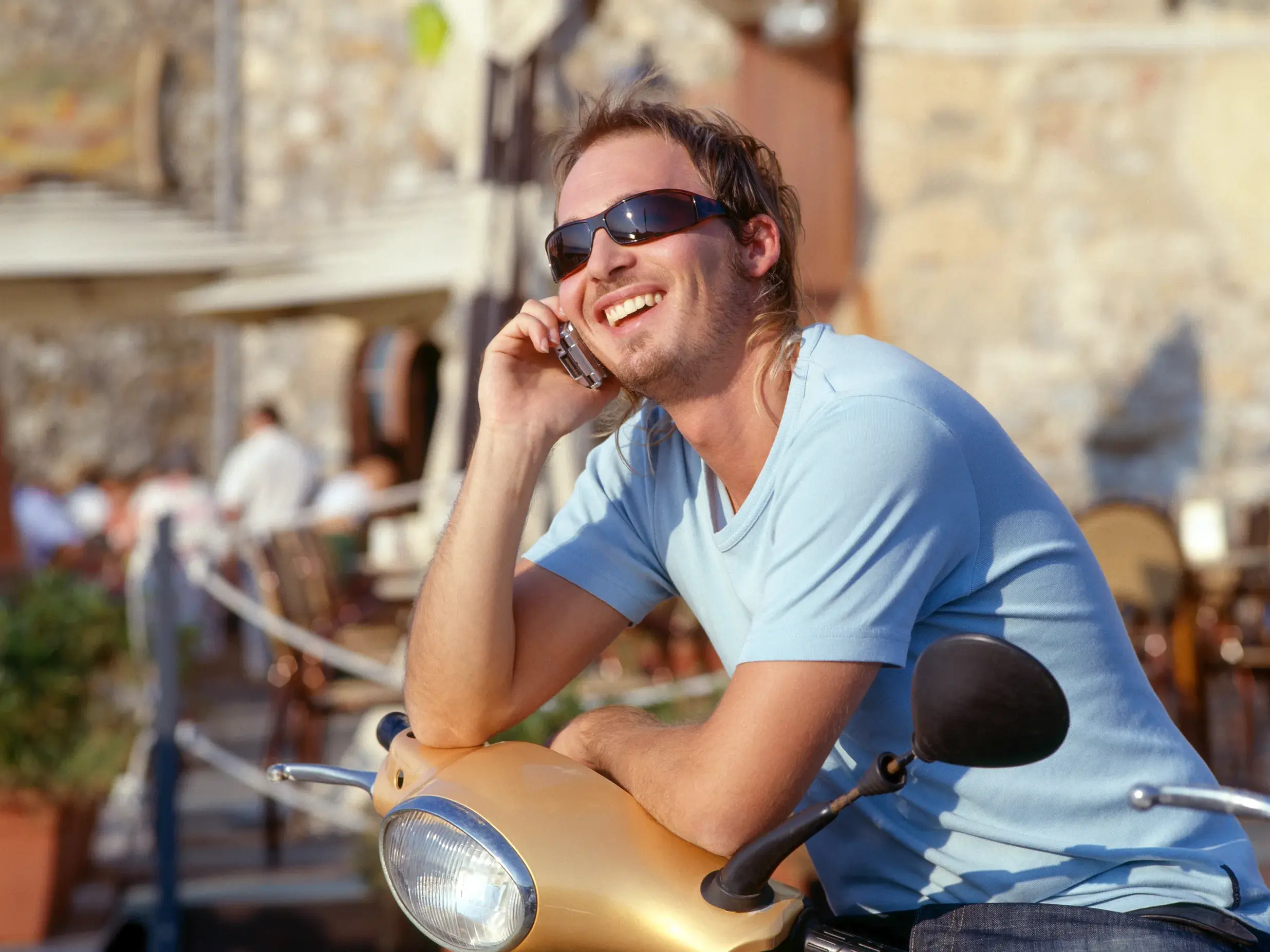 Ein Mann mit Sonnenbrille telefoniert lächelnd auf seinem Motorroller in einer sonnigen, belebten Gasse in Neapel.