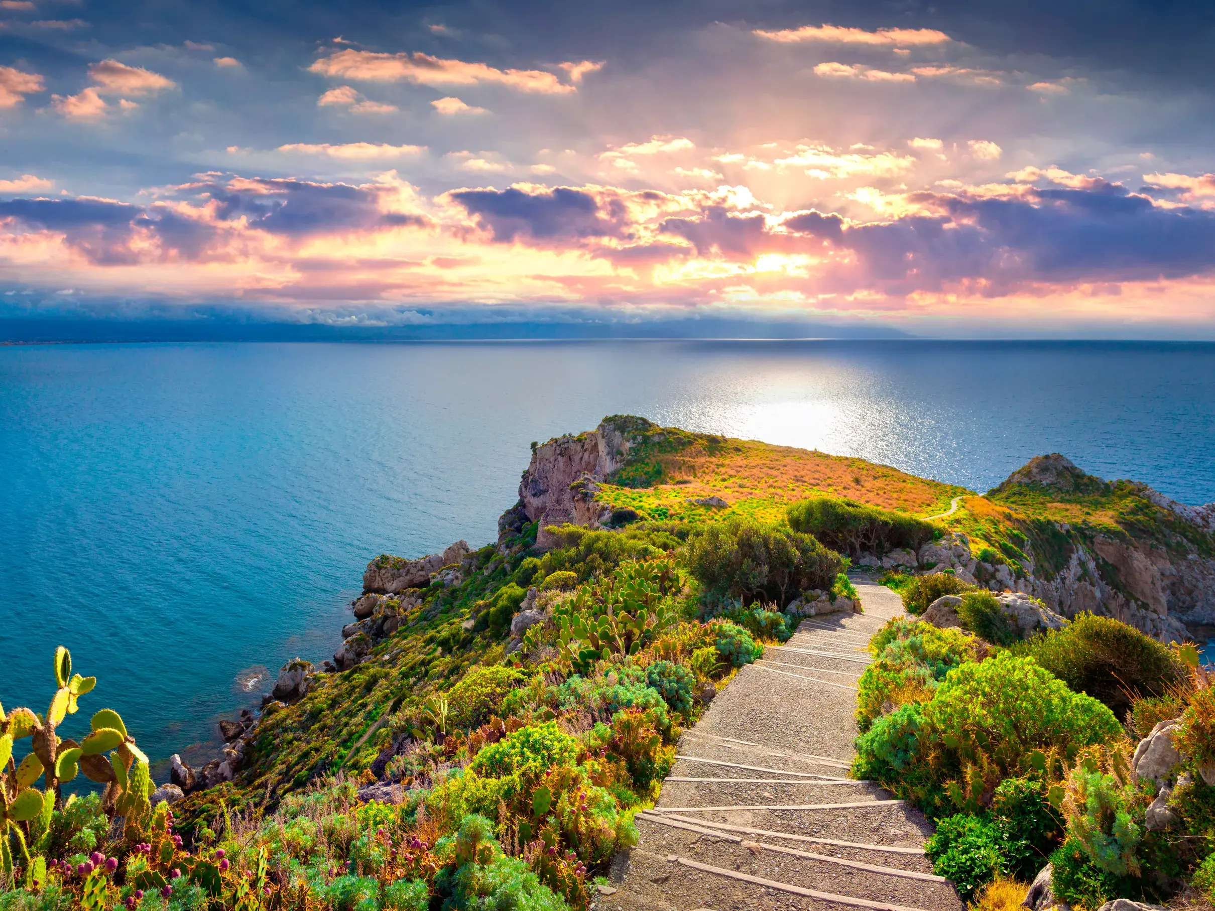 Steiniger Pfad führt entlang der grünen Steilküste in Sizilien hinab zum Meer bei Sonnenuntergang.