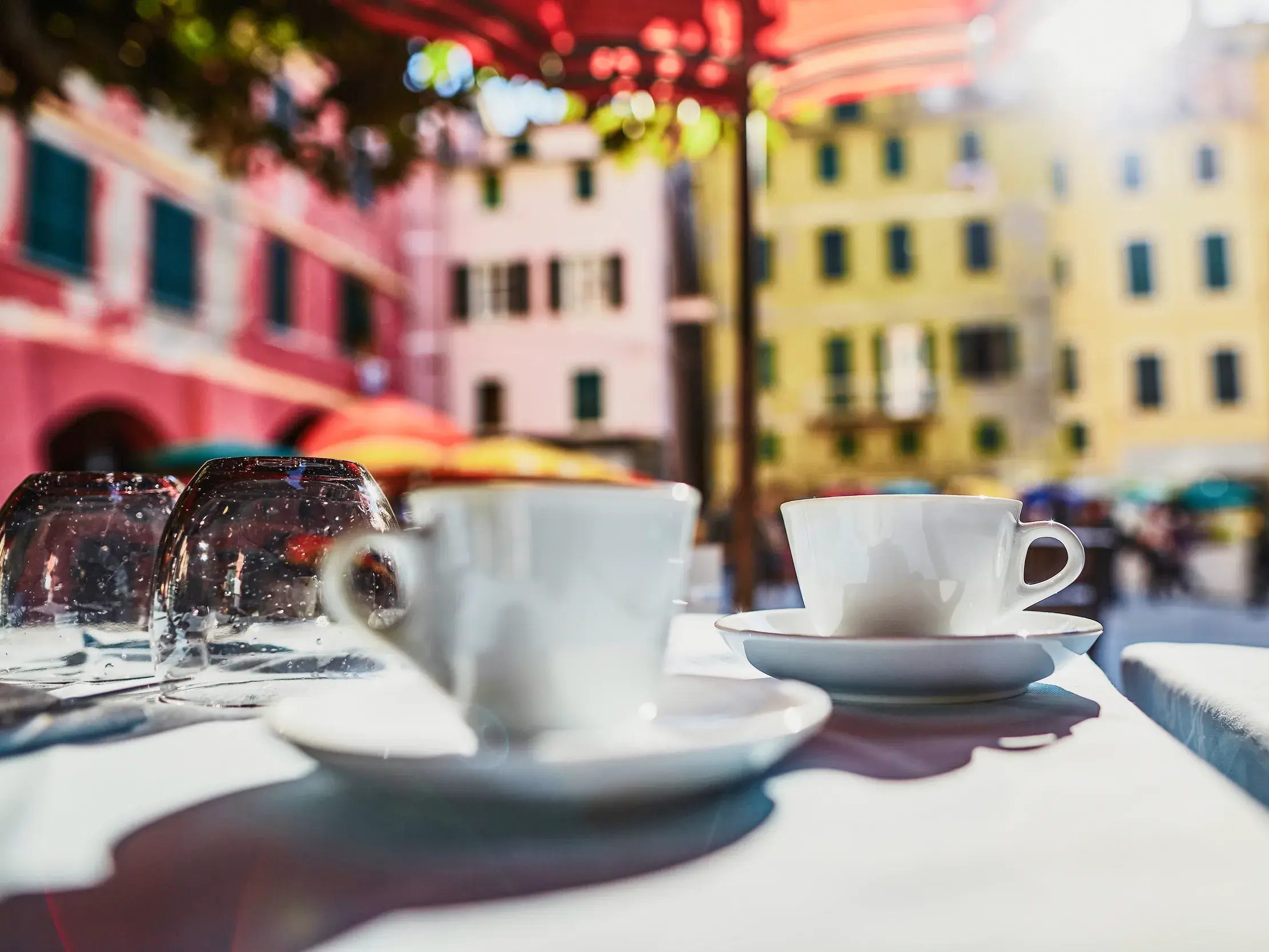 Zwei Kaffeetassen auf einem gedeckten Tisch vor der sonnigen, bunten Häuserkulisse von Vernazza in der Cinqueterre.