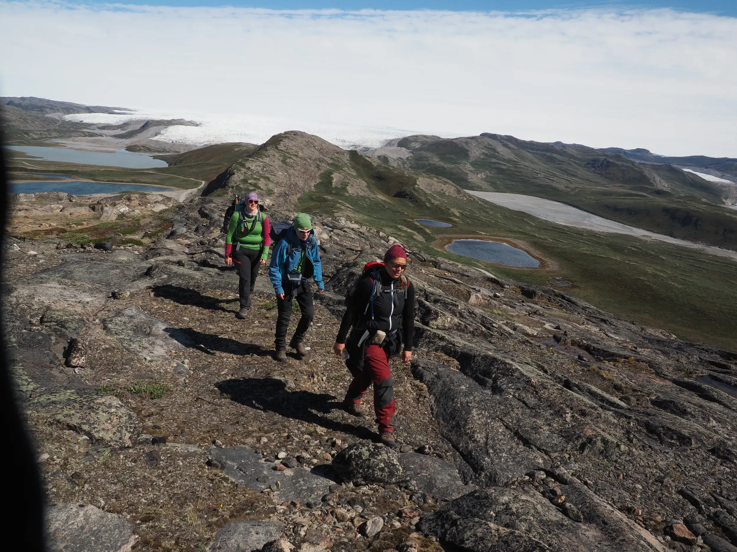 Eine kleine Wandergruppe beim Trekking auf einem felsigen Höhenzug in Grönland mit Panoramablick auf das weite Inlandeis.
