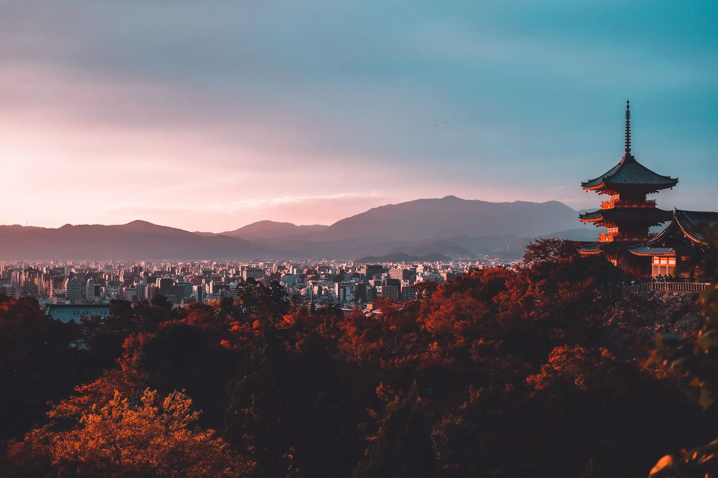 Eine rote Pagode thront über dem Herbstlaub mit Panoramablick auf die Stadt Kyoto und die Berge in der Abenddämmerung.