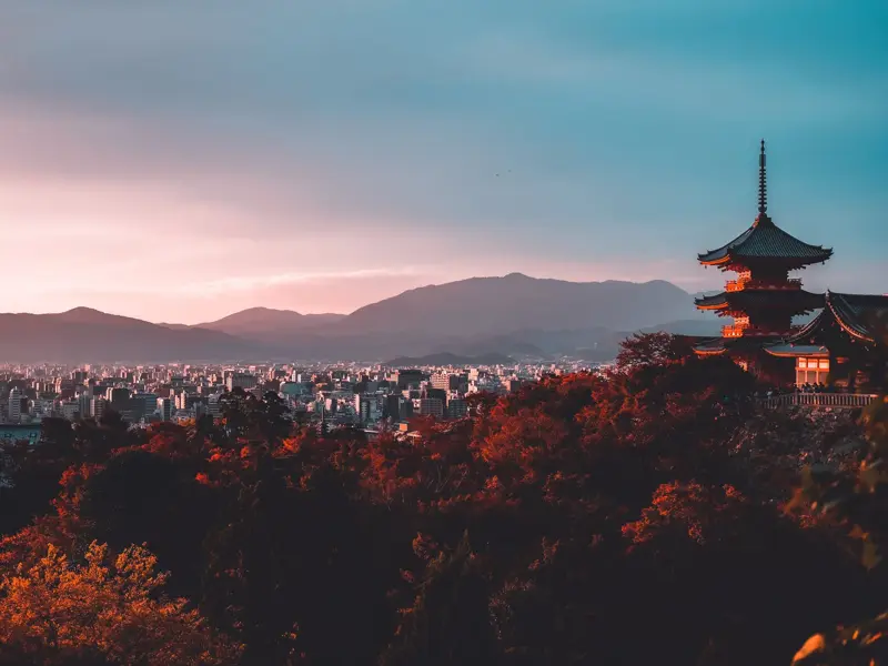 Eine rote Pagode thront über dem Herbstlaub mit Panoramablick auf die Stadt Kyoto und die Berge in der Abenddämmerung.