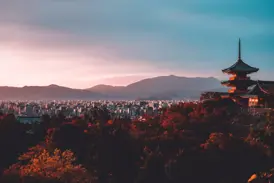 Eine rote Pagode thront über dem Herbstlaub mit Panoramablick auf die Stadt Kyoto und die Berge in der Abenddämmerung.