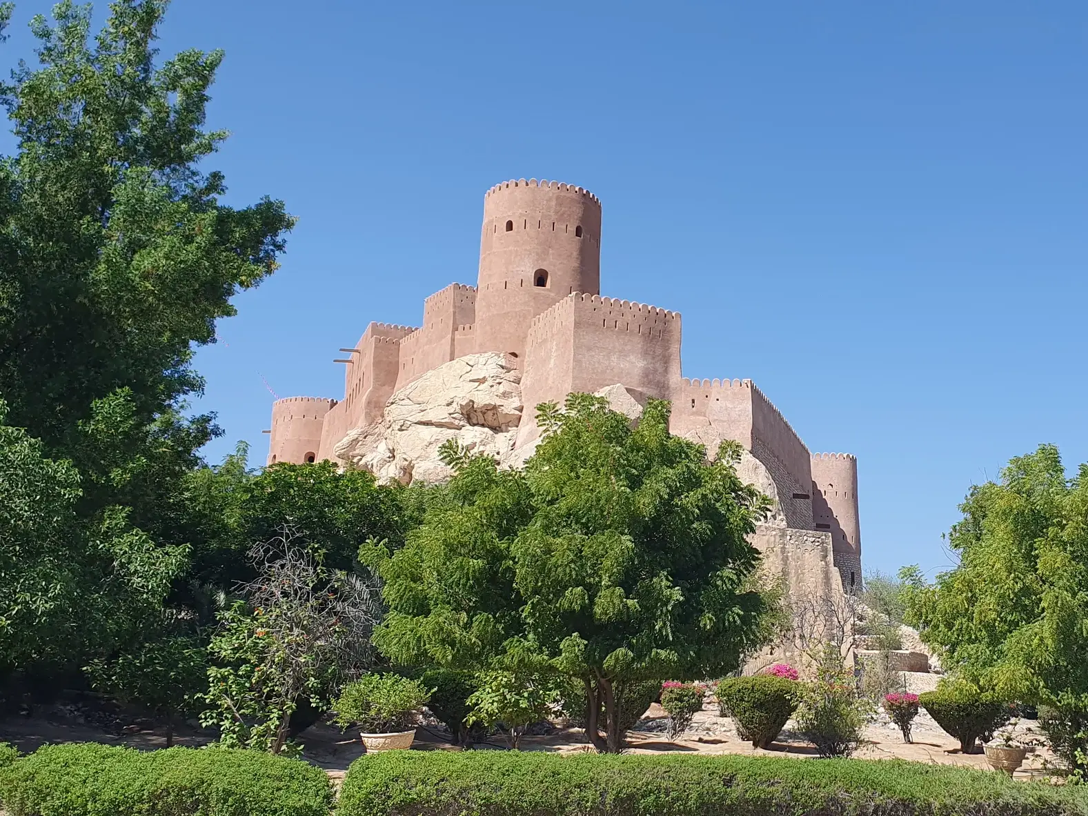 Die lehmfarbene Festung von Nakhl im Oman thront auf einem Felsen über einem grünen Palmenhain bei Sonnenschein.