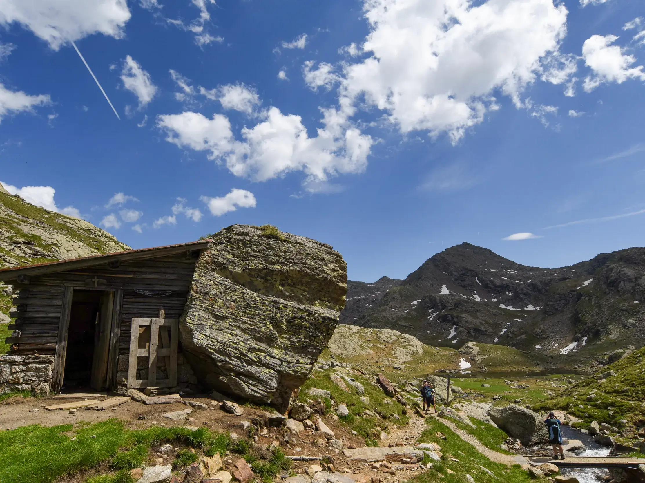 Rustikale Holzhütte in der alpinen Felslandschaft der Texelgruppe in Südtirol, im Hintergrund wandern zwei Personen auf einem Pfad.