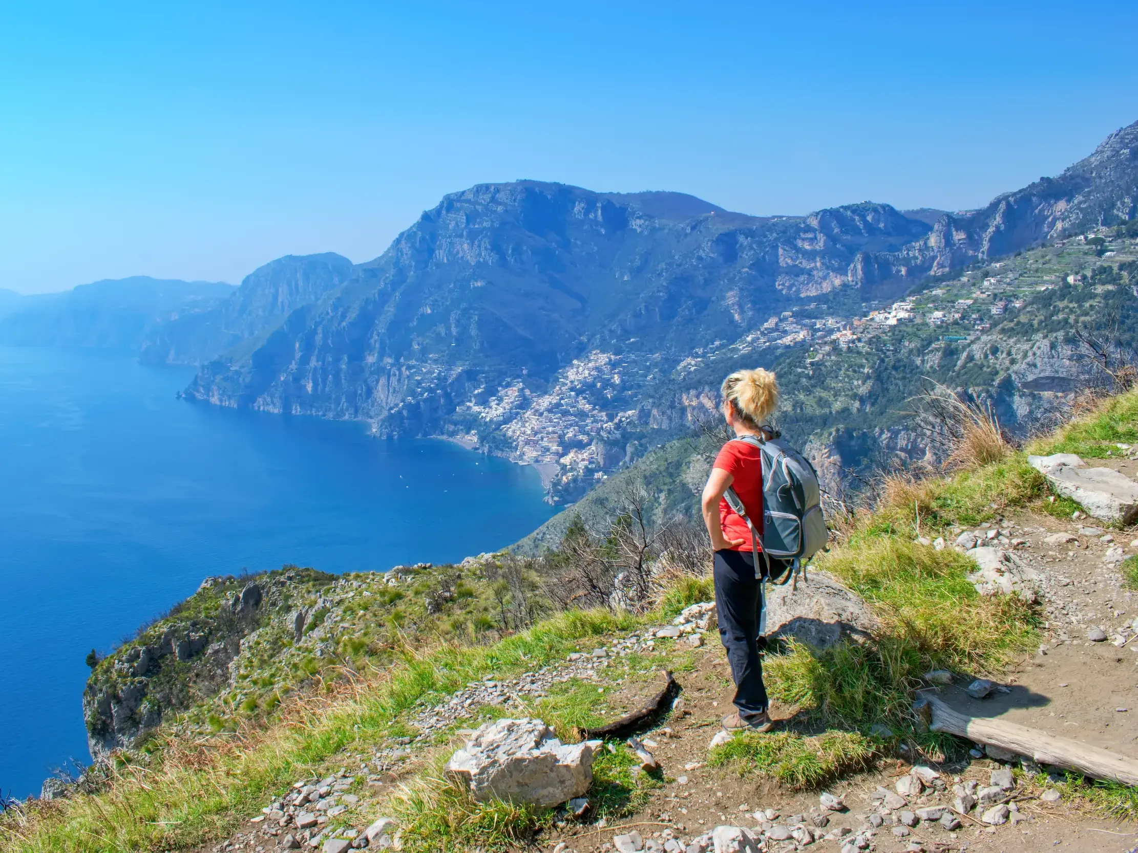 Eine Wanderin pausiert auf dem Sentiero degli Dei und blickt auf die steilen Klippen und das blaue Meer der Amalfiküste.