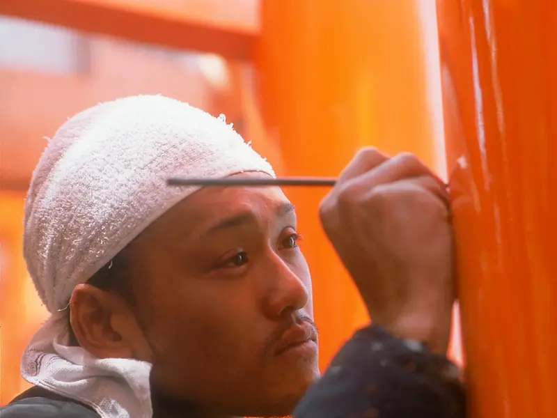 Ein japanischer Handwerker mit weißem Stirnband restauriert konzentriert die leuchtend orange Lackierung eines Torii-Tors in Japan.