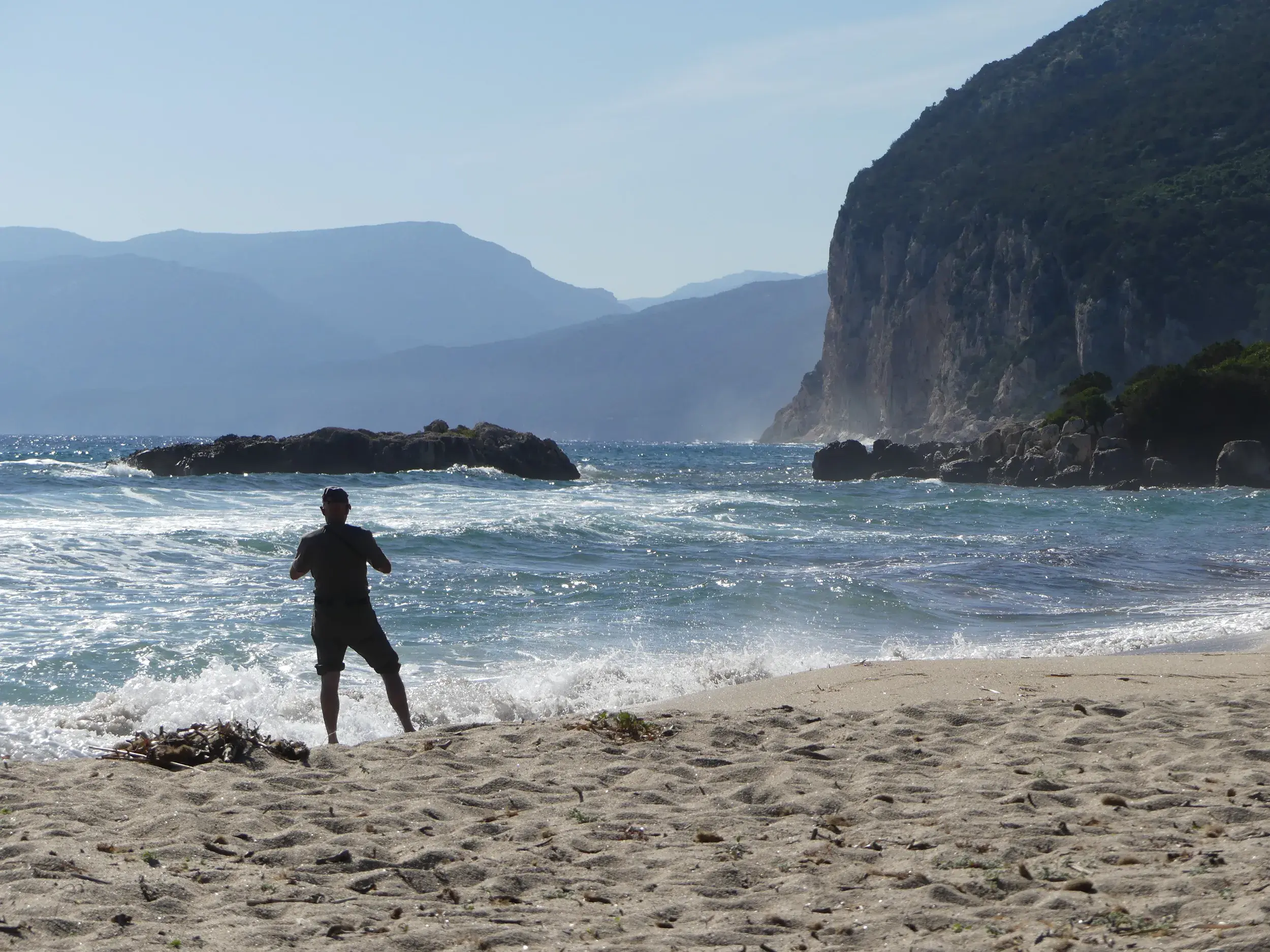 Ein Wanderer steht barfuß an einem Sandstrand auf Sardinien, blickt auf die Wellen und die felsige Steilküste am Horizont.