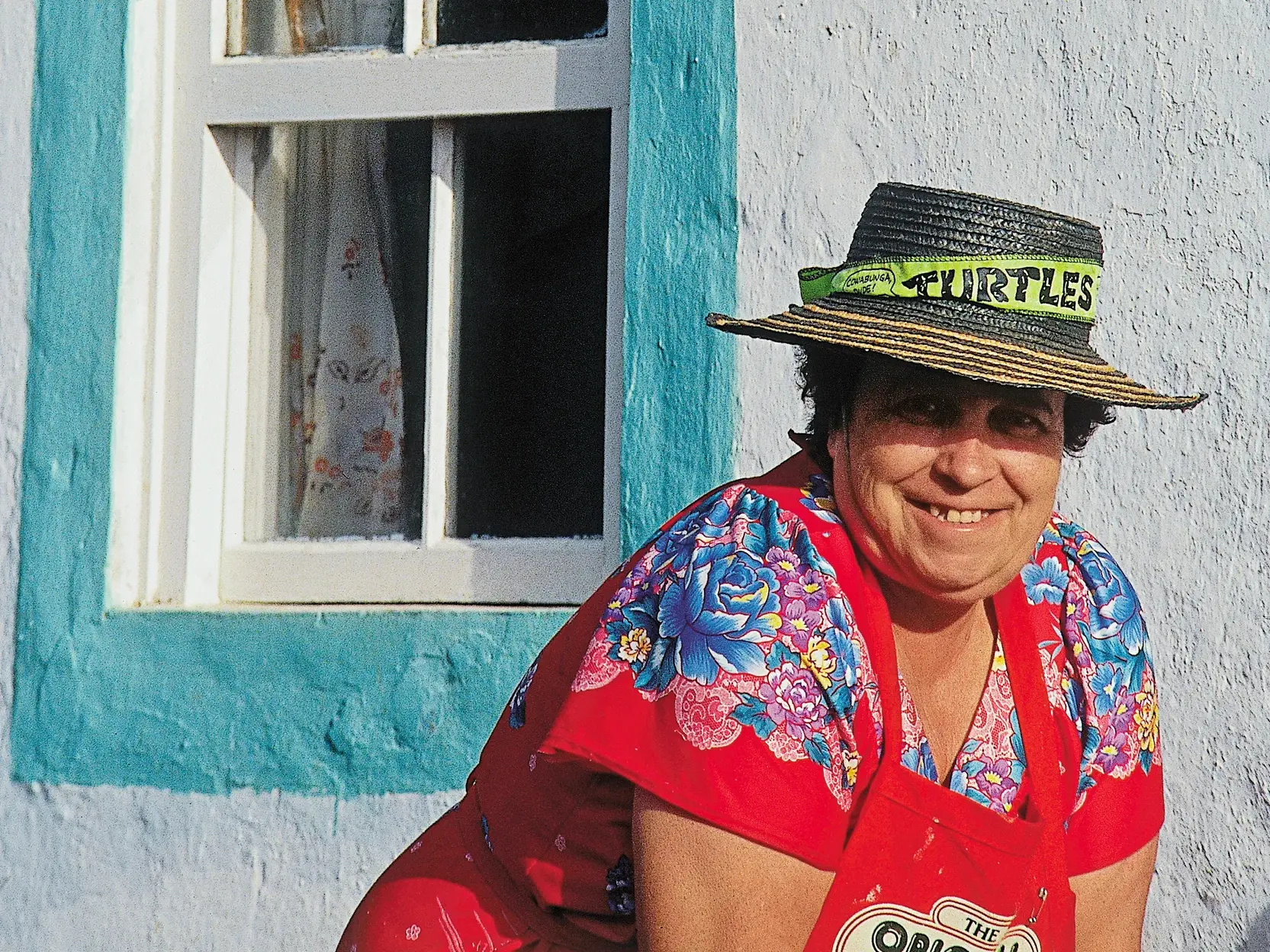 Eine lächelnde Frau mit Strohhut sitzt vor der bunten Hausfassade in Angra do Heroísmo auf der Insel Terceira.