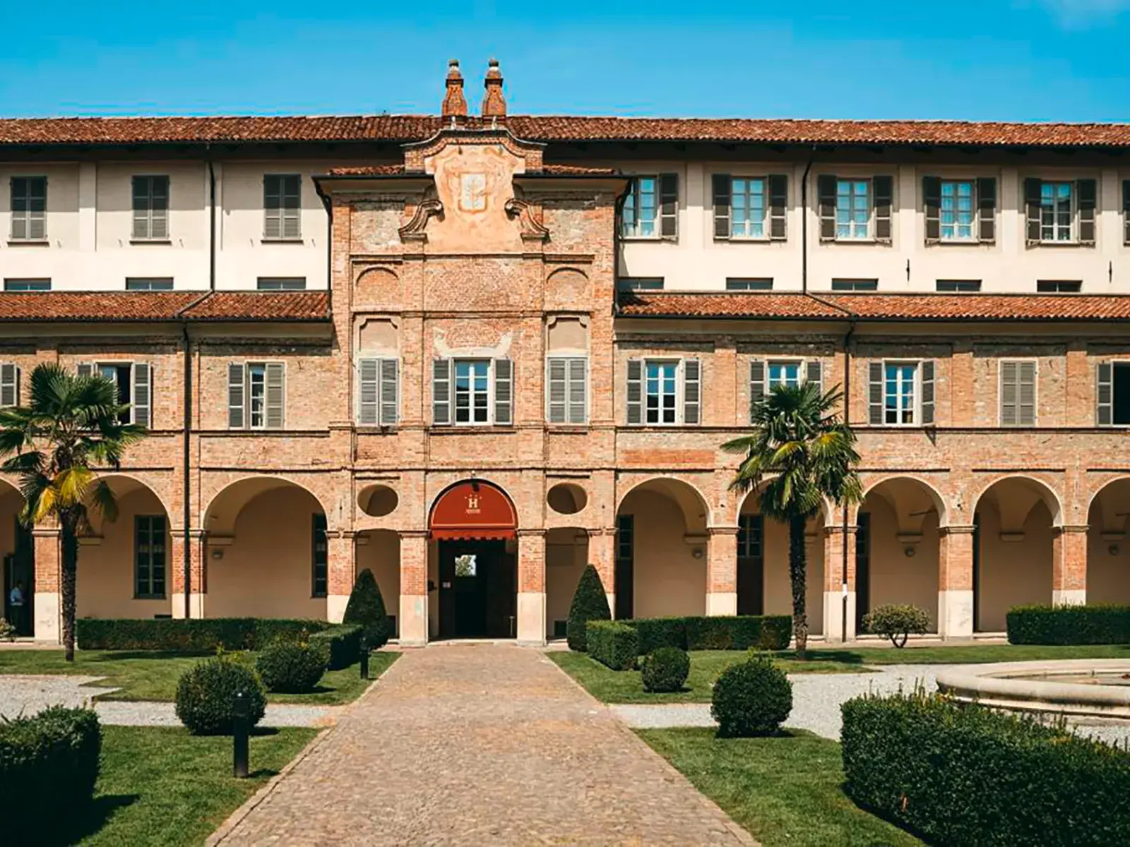 Die historische Fassade des Hotels I Somaschi in Cherasco mit dem gepflegten Innenhof und Palmen bei Sonnenschein.