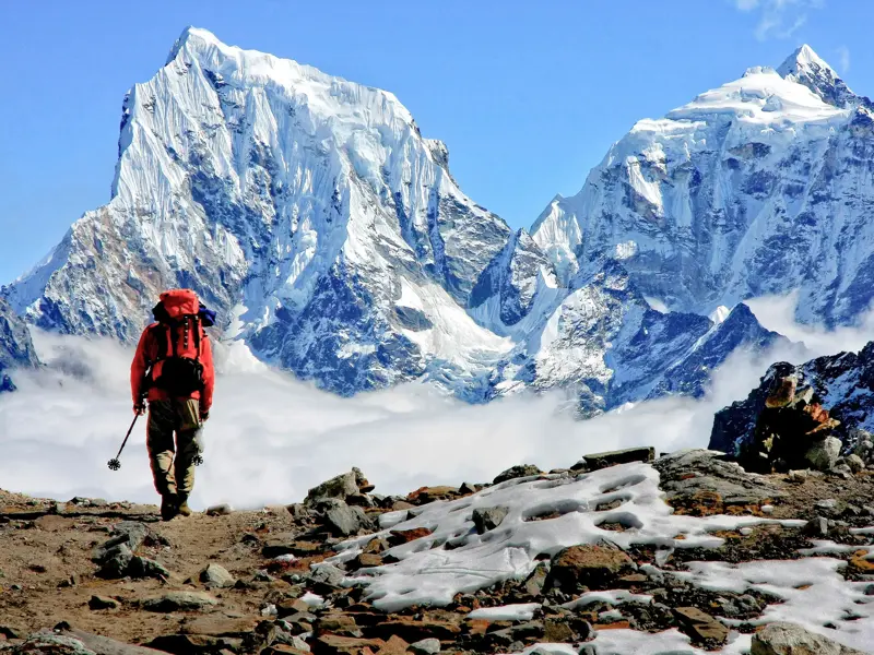 Trekker mit rotem Rucksack auf einem steinigen Bergpfad vor der majestätischen, schneebedeckten Ama Dablam in Nepal.