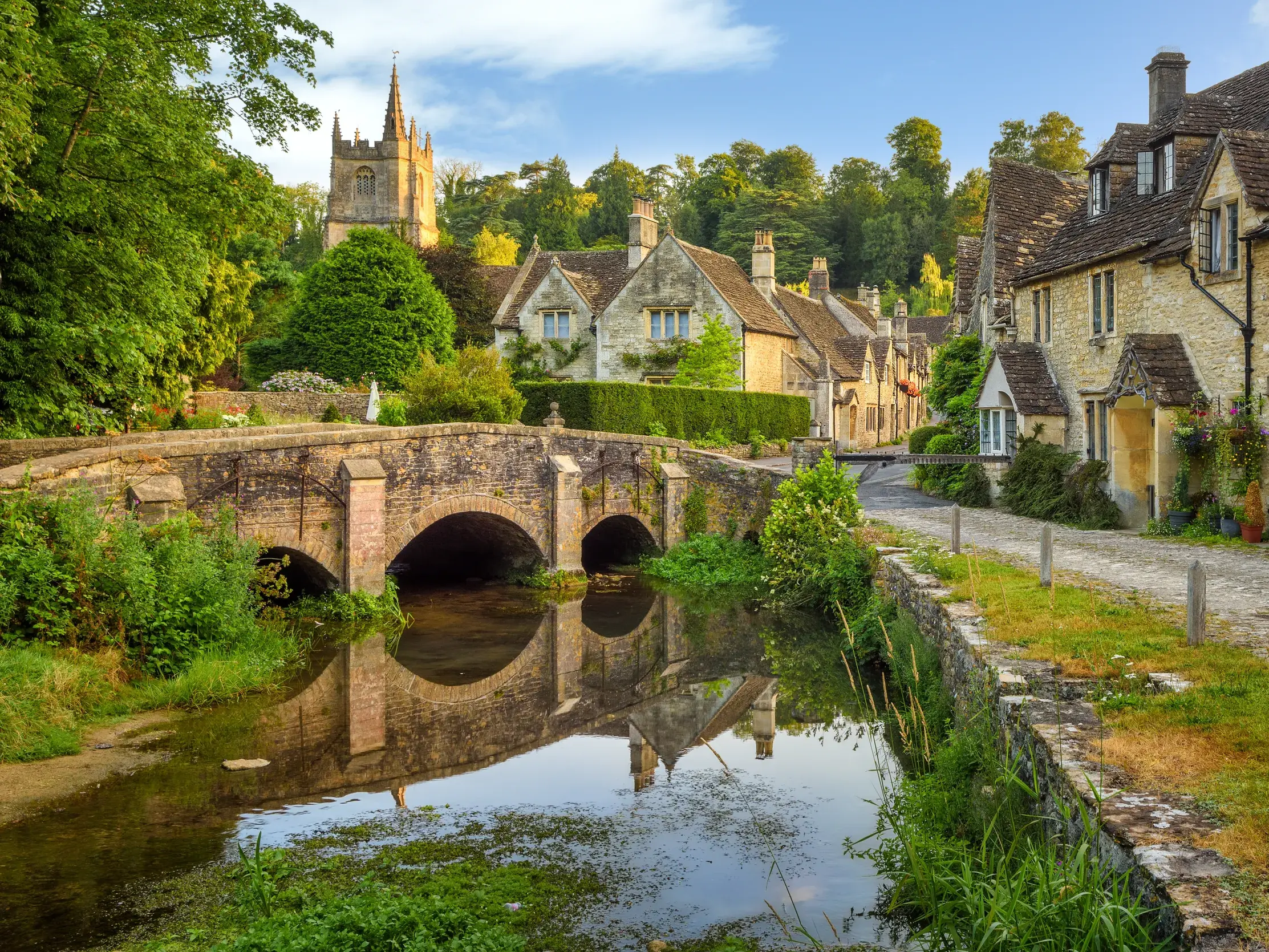 Idyllisches Bilderbuchdorf in den Cotswolds, Südengland: Eine alte Steinbrücke spiegelt sich in einem Fluss vor honigfarbenen Cottages.