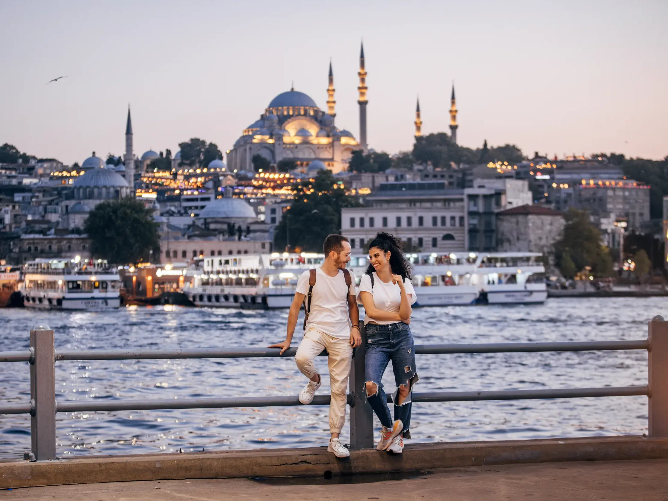 Junges Paar lehnt am Geländer am Bosporus mit Blick auf die beleuchtete Süleyman-Moschee in Istanbul bei Dämmerung.