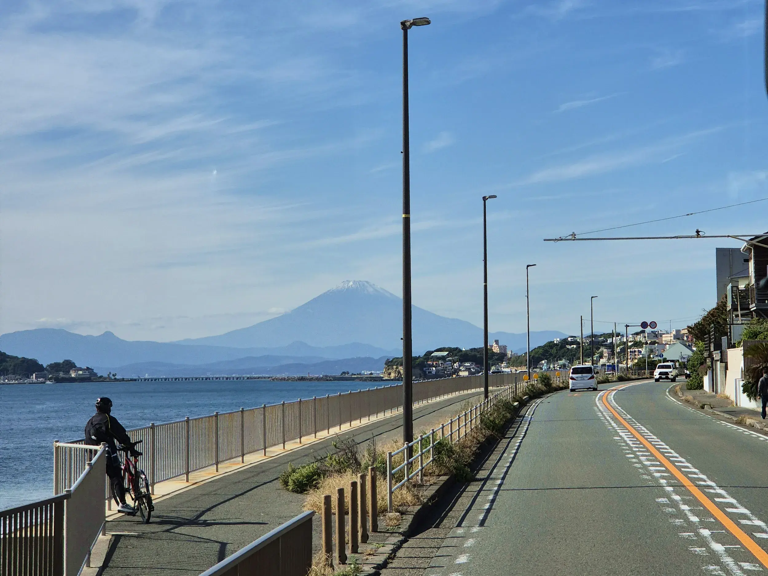 Radfahrer an einer Küstenpromenade bei Kamakura genießt bei Sonnenschein den weiten Blick über die Bucht auf den Mount Fuji.