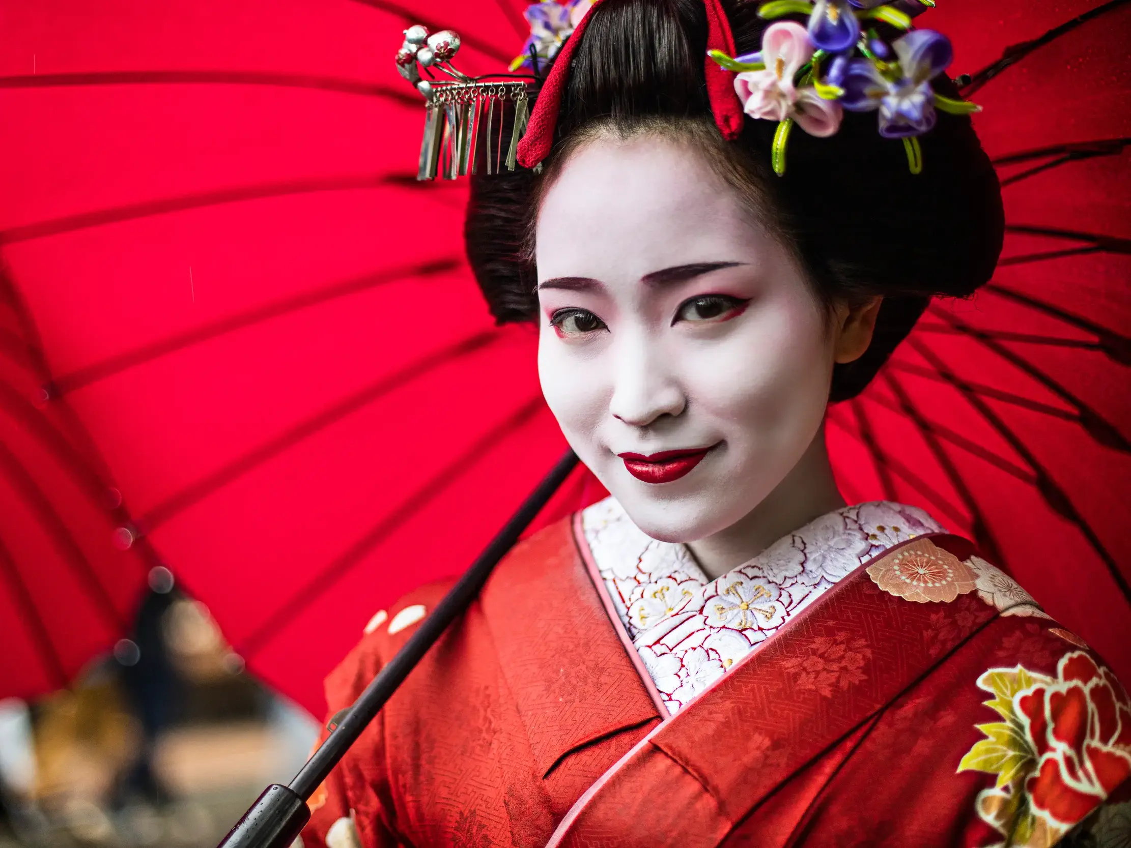 Traditionell geschminkte Geisha in rotem Kimono mit kunstvollem Haarschmuck lächelt unter einem roten Papierschirm in Kyoto.