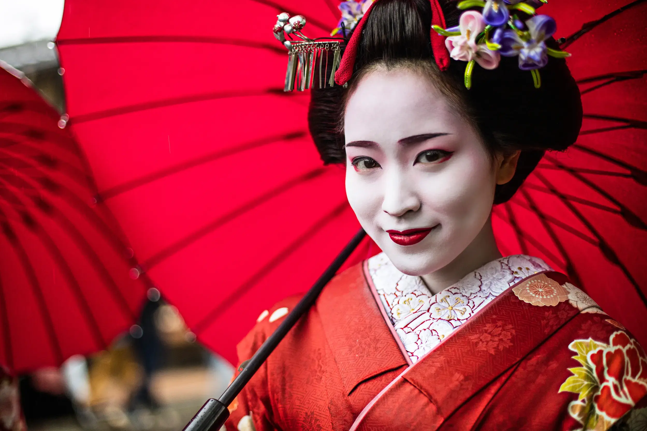 Traditionell geschminkte Geisha in rotem Kimono mit kunstvollem Haarschmuck lächelt unter einem roten Papierschirm in Kyoto.