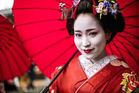 Traditionell geschminkte Geisha in rotem Kimono mit kunstvollem Haarschmuck lächelt unter einem roten Papierschirm in Kyoto.