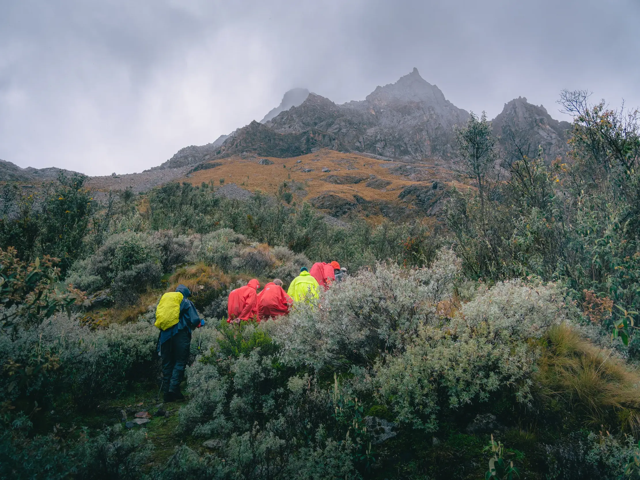 Eine Wandergruppe in Regenjacken wandert auf einem schmalen Pfad durch die raue Berglandschaft der Cordillera Blanca in Peru.