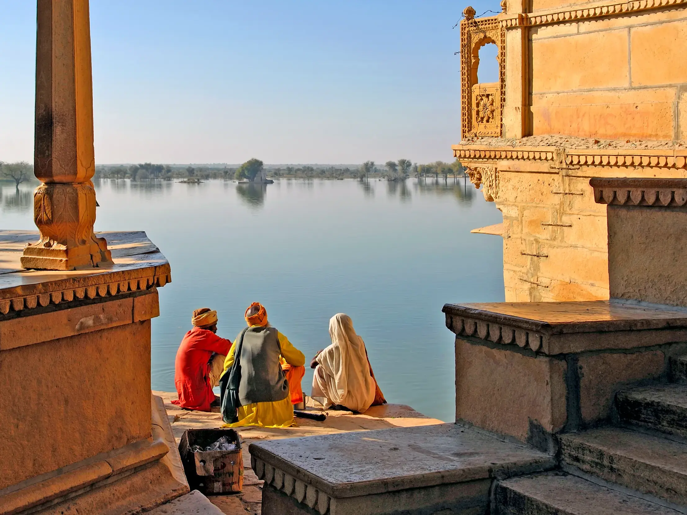Rückansicht von drei Einheimischen in bunten Gewändern auf einer Steinterrasse am Ufer des Gadisar Sees in Jaisalmer, Rajasthan.