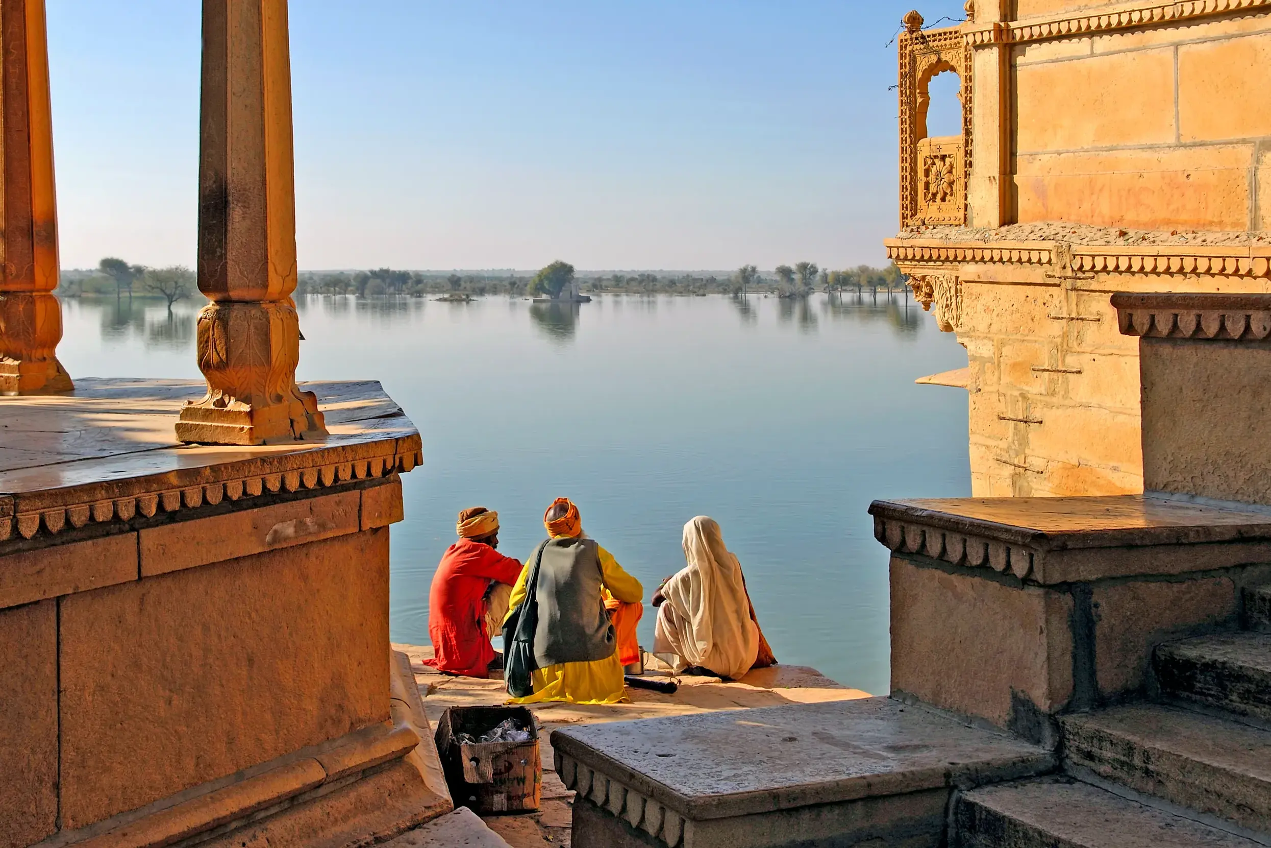 Rückansicht von drei Einheimischen in bunten Gewändern auf einer Steinterrasse am Ufer des Gadisar Sees in Jaisalmer, Rajasthan.