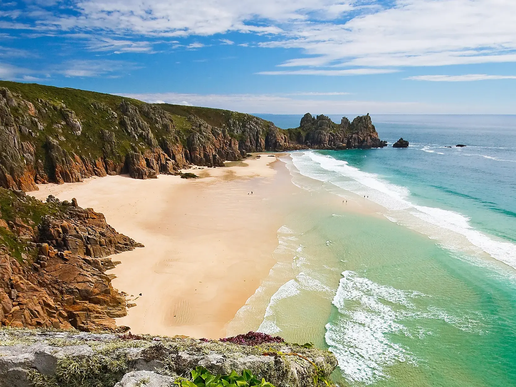 Erhöhter Blick auf die wilde Granitküste bei Land's End in Cornwall, mit Sandstrand und türkisfarbenem Meer bei Sonnenschein.