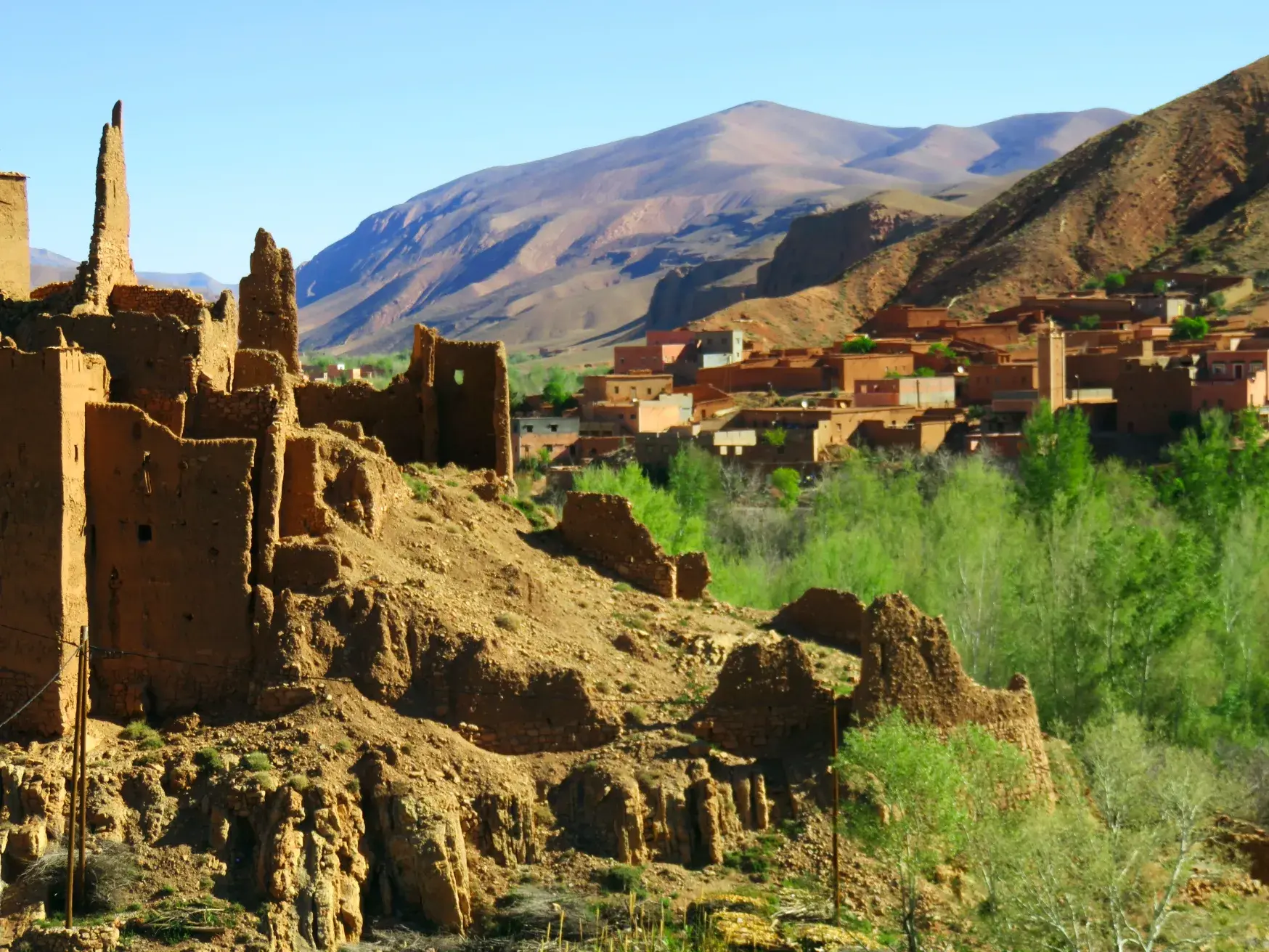 Ruinen einer Lehm-Kasbah an einem grünen Flusstal, im Hintergrund ein Berberdorf vor der Bergkulisse des Hohen Atlas.