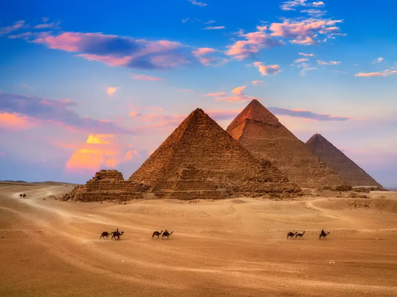 Die drei großen Pyramiden von Gizeh in der ägyptischen Wüste bei Sonnenuntergang. Eine kleine Karawane reitet auf Kamelen.