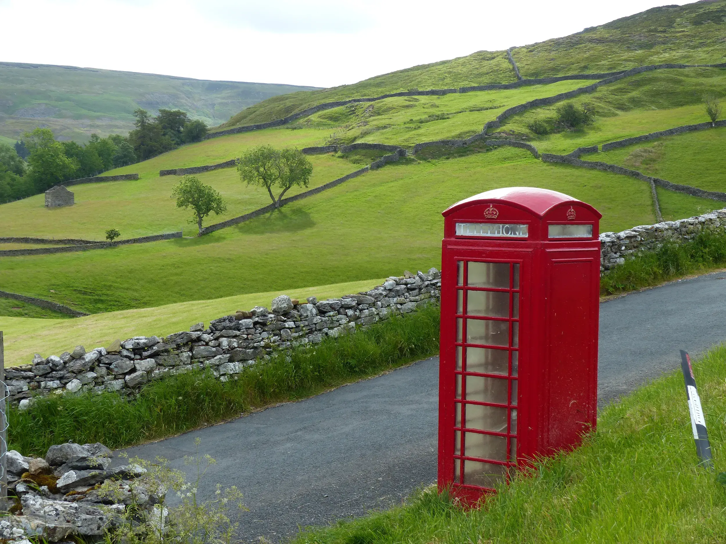 Rote Telefonzelle an einer schmalen Straße inmitten der weiten, grünen Hügellandschaft der Yorkshire Dales mit Steinmauern.