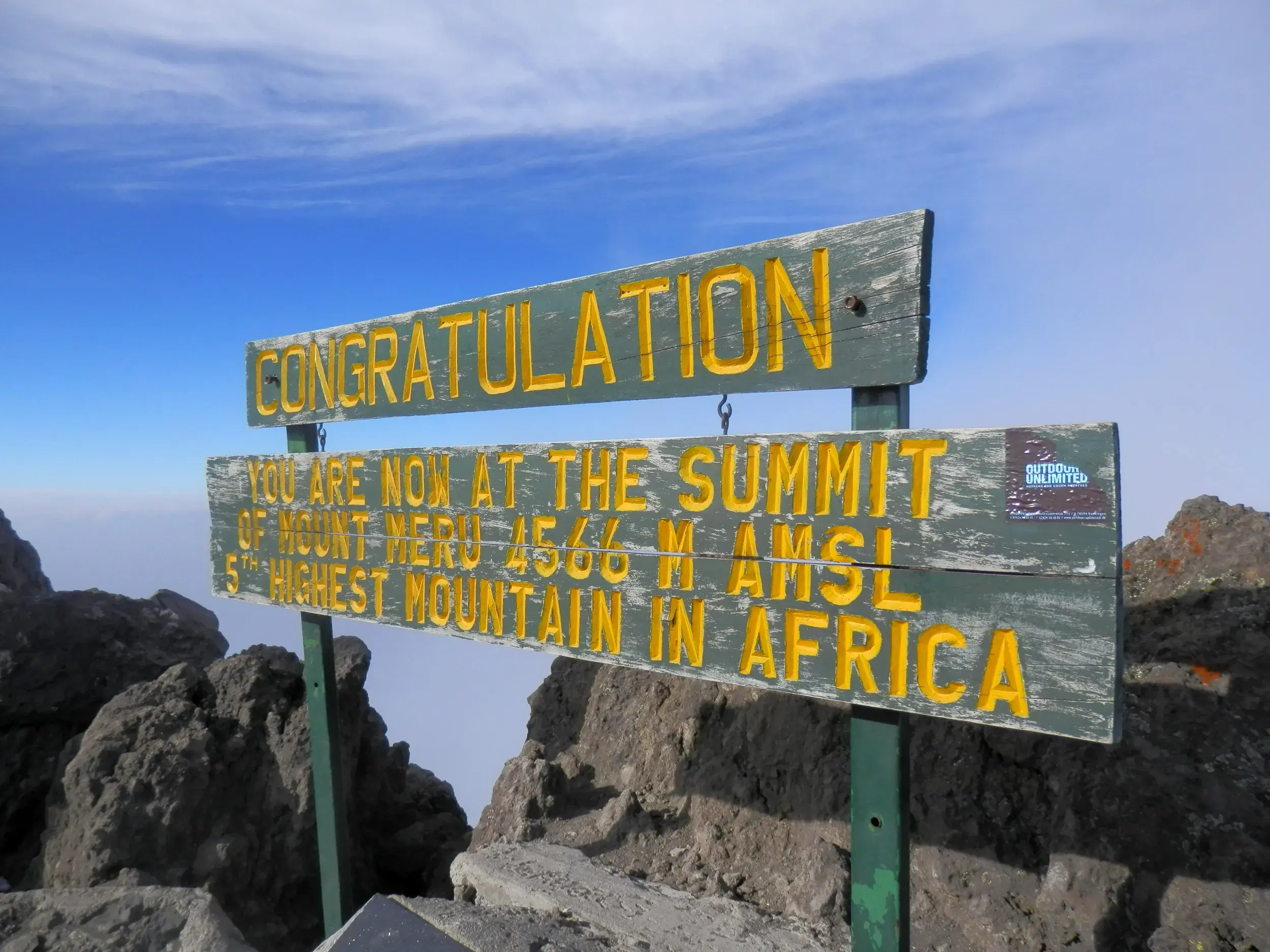 Das hölzerne Gipfelschild auf dem 4.566 m hohen Mount Meru in Tansania, das über einem dichten Wolkenmeer thront.