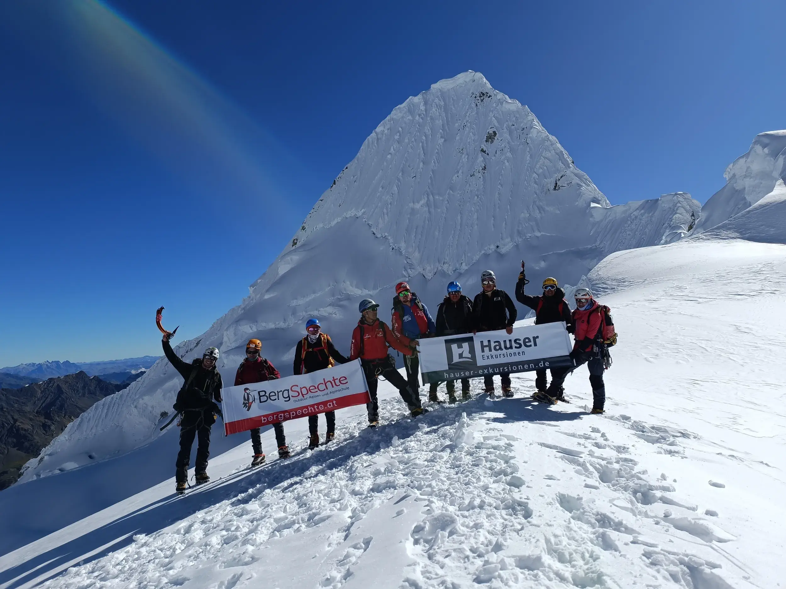 Glückliche Bergsteiger-Gruppe mit Hauser Exkursionen Fahne auf einem Gipfelgrat in Peru mit Blick auf den schneebedeckten Alpamayo.