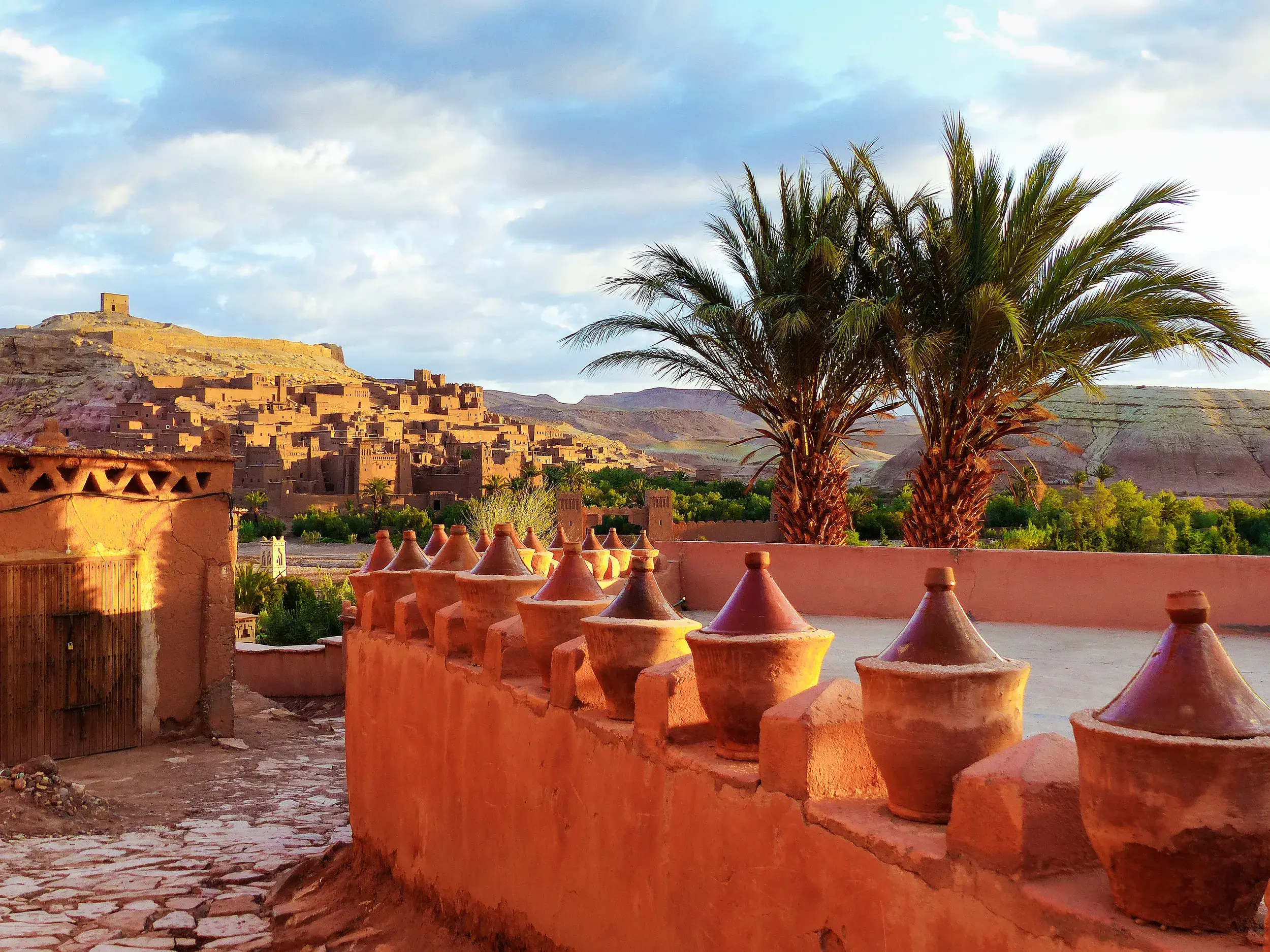 Die historischen Lehmbauten der Kasbah Aït Benhaddou in Marokko, eingerahmt von Palmen und einer traditionellen Lehm-Mauer.