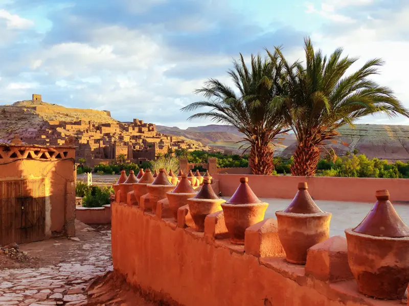 Die historischen Lehmbauten der Kasbah Aït Benhaddou in Marokko, eingerahmt von Palmen und einer traditionellen Lehm-Mauer.