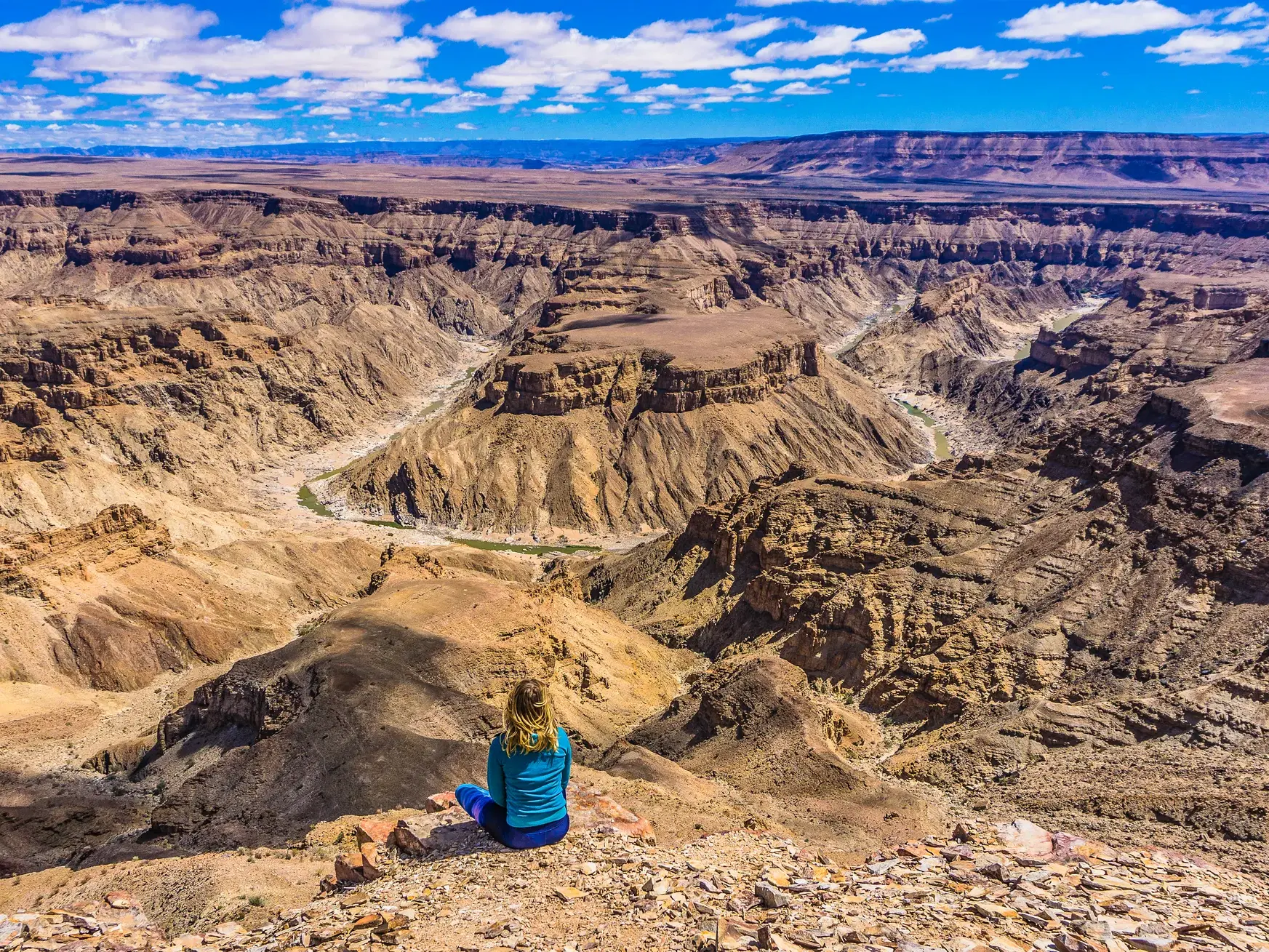 Eine Reisende sitzt am Rande des gewaltigen Fish River Canyons in Namibia und blickt über die weite, sonnige Schlucht.