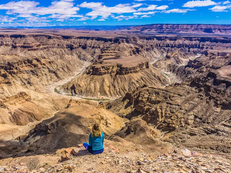 Eine Reisende sitzt am Rande des gewaltigen Fish River Canyons in Namibia und blickt über die weite, sonnige Schlucht.