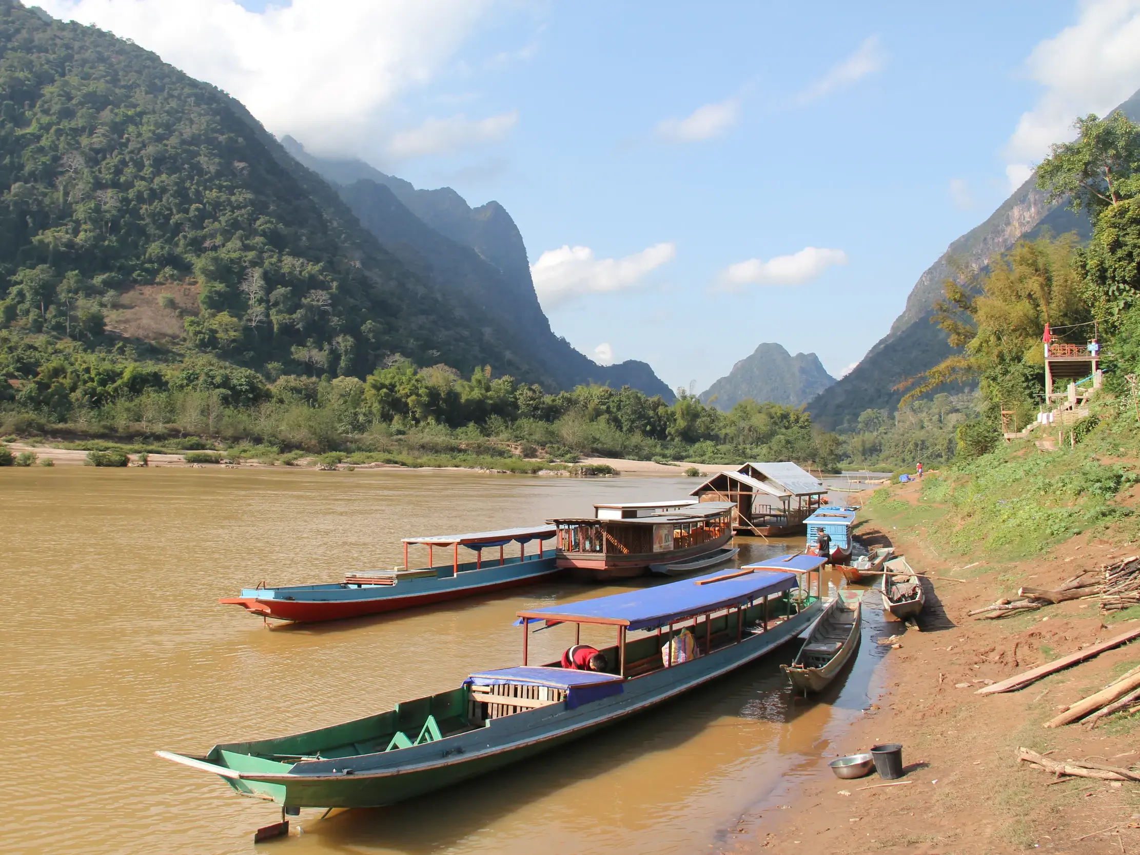 Traditionelle Longtail-Boote ankern am Ufer des Nam Ou Flusses in Laos, umgeben von der malerischen Kulisse grüner Karstberge.