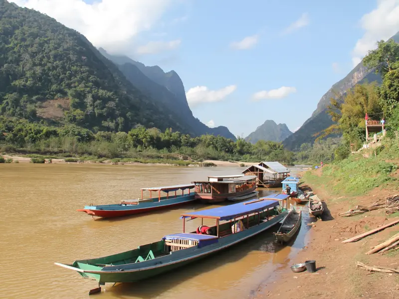Traditionelle Longtail-Boote ankern am Ufer des Nam Ou Flusses in Laos, umgeben von der malerischen Kulisse grüner Karstberge.