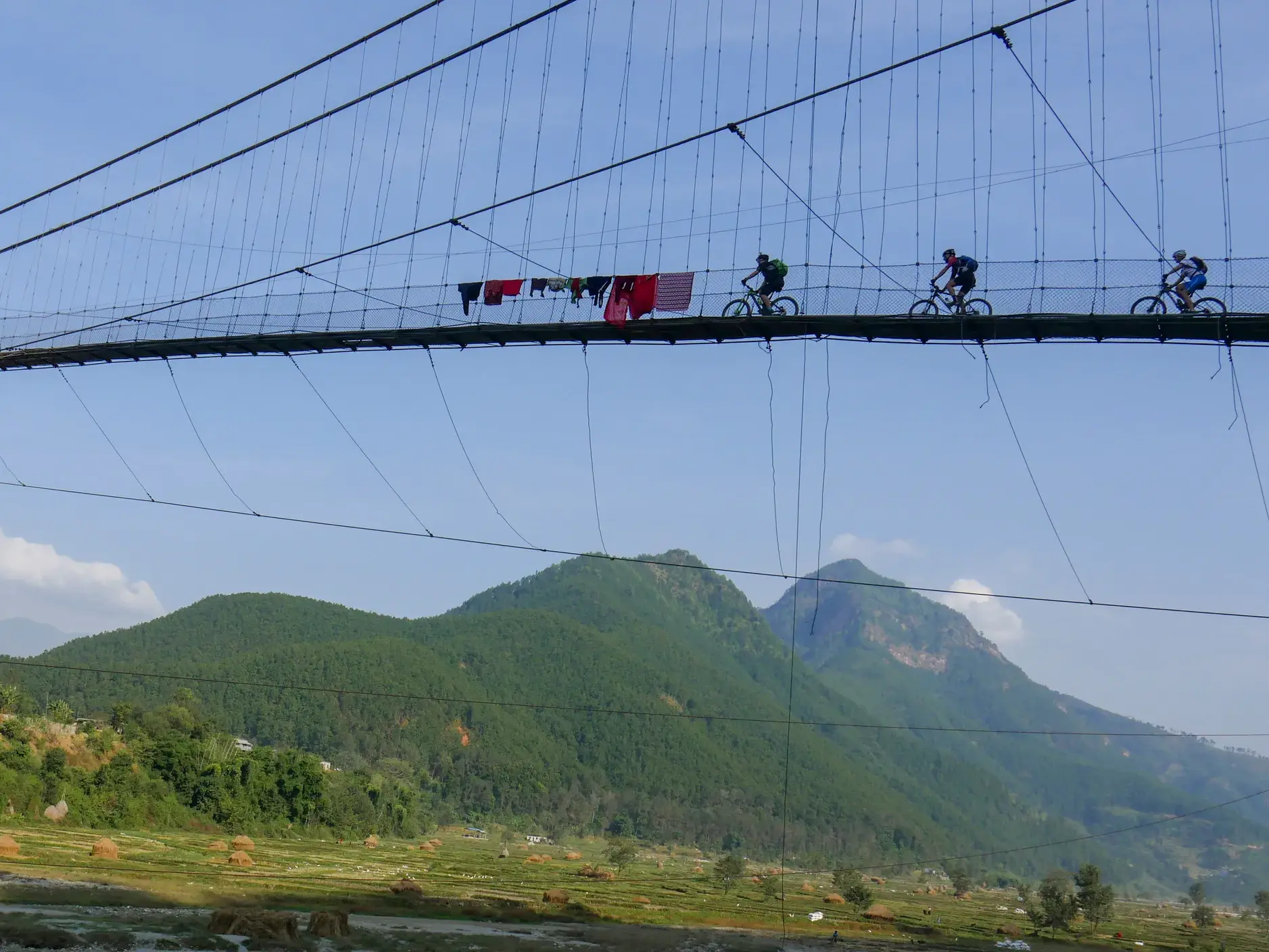 Eine Gruppe Mountainbiker überquert eine hohe Hängebrücke in Nepal, im Hintergrund die grünen Ausläufer des Himalaya.