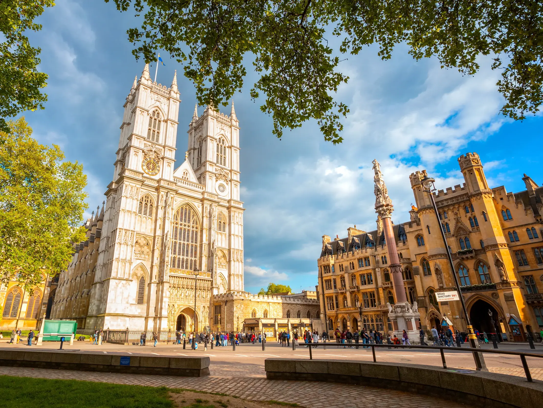 Gotische Pracht der Westminster Abbey in London, gesehen von einem belebten Platz mit Touristen und umrahmt von grünen Blättern.