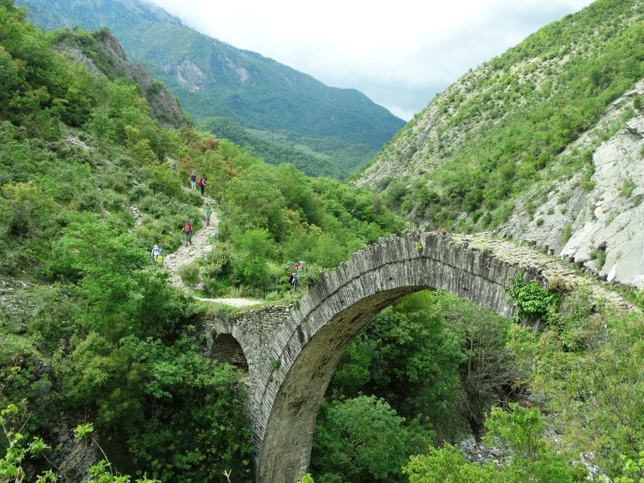 Eine Wandergruppe überquert auf einem Trekkingpfad eine alte osmanische Steinbrücke im grünen Zagoria-Tal in Albanien.
