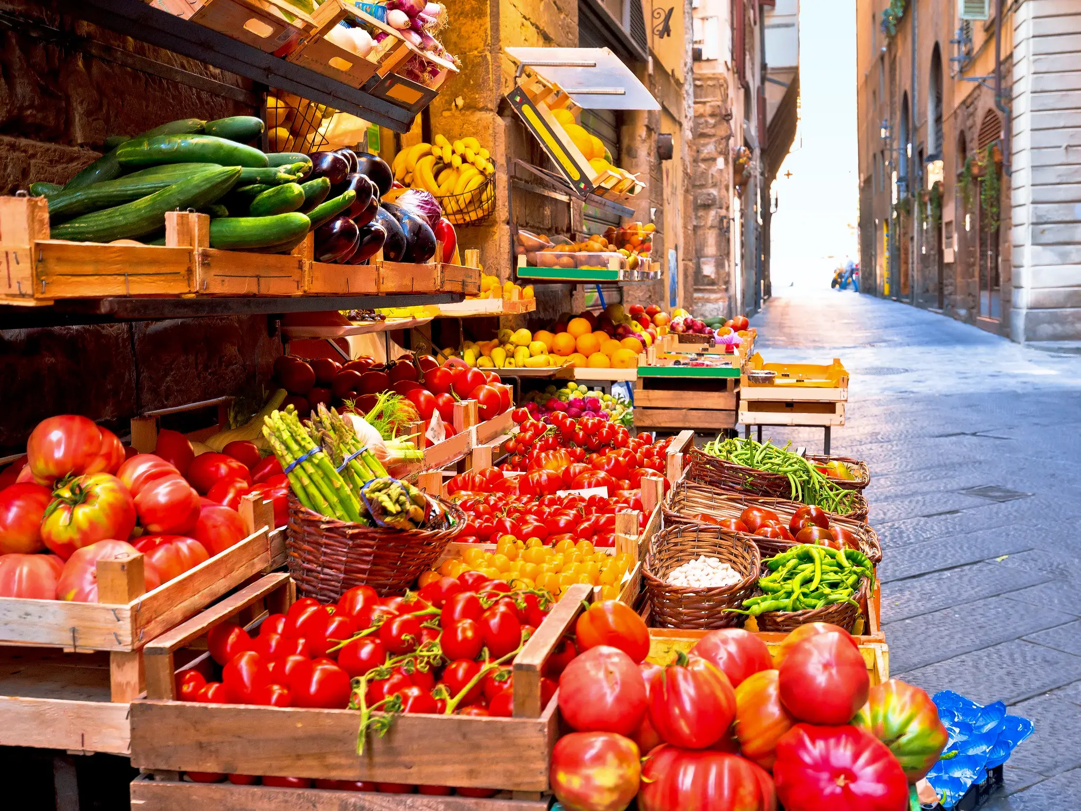 Bunter Marktstand mit frischem Gemüse wie Tomaten und Spargel in Holzkisten in einer engen Gasse in Florenz.