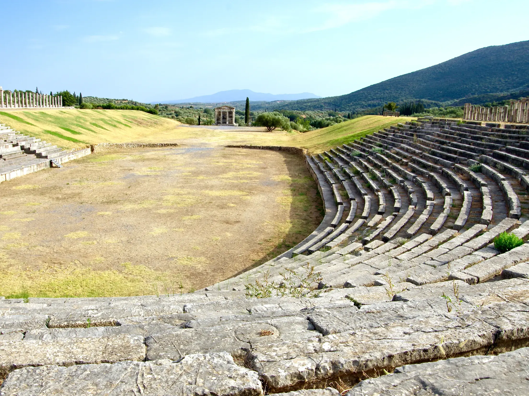 Antikes Stadion von Messene mit Steintribünen und Blick auf die grüne Hügellandschaft des Peloponnes an einem sonnigen Tag.