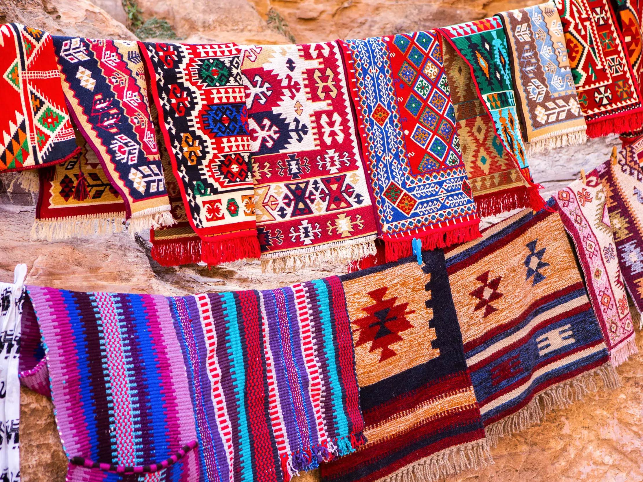 Farbenfrohe, handgewebte Teppiche mit Beduinenmustern hängen als Souvenirs an einer Leine vor einer Felswand in Petra.