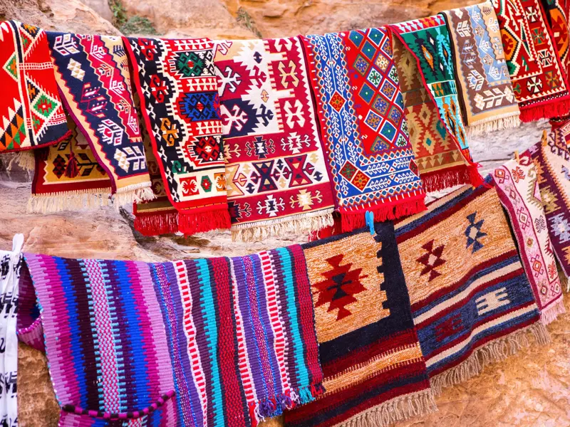 Farbenfrohe, handgewebte Teppiche mit Beduinenmustern hängen als Souvenirs an einer Leine vor einer Felswand in Petra.