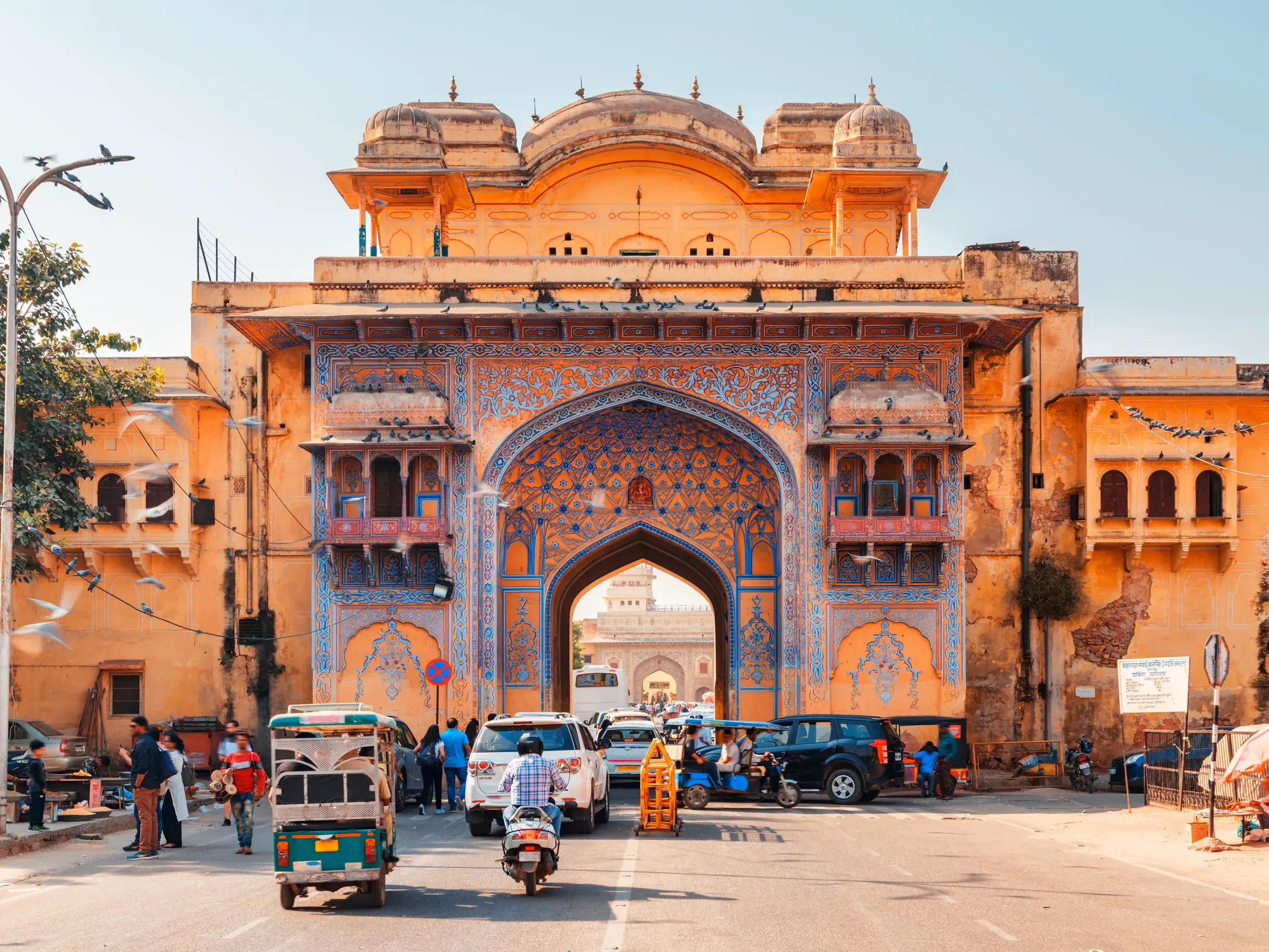 Lebhafter Straßenverkehr mit Tuk-Tuks vor einem reich verzierten, historischen Stadttor in der rosaroten Stadt Jaipur, Indien.
