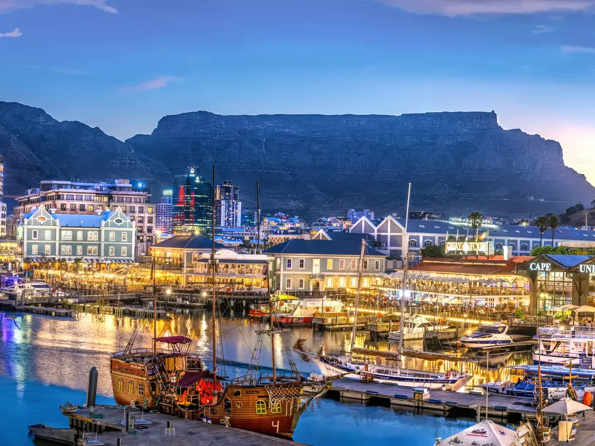 Die belebte V&A Waterfront in Kapstadt mit Restaurants und Booten im Hafen vor der Kulisse des Tafelbergs bei Dämmerung besuchen die Gäste der Studiosus Studienreise.