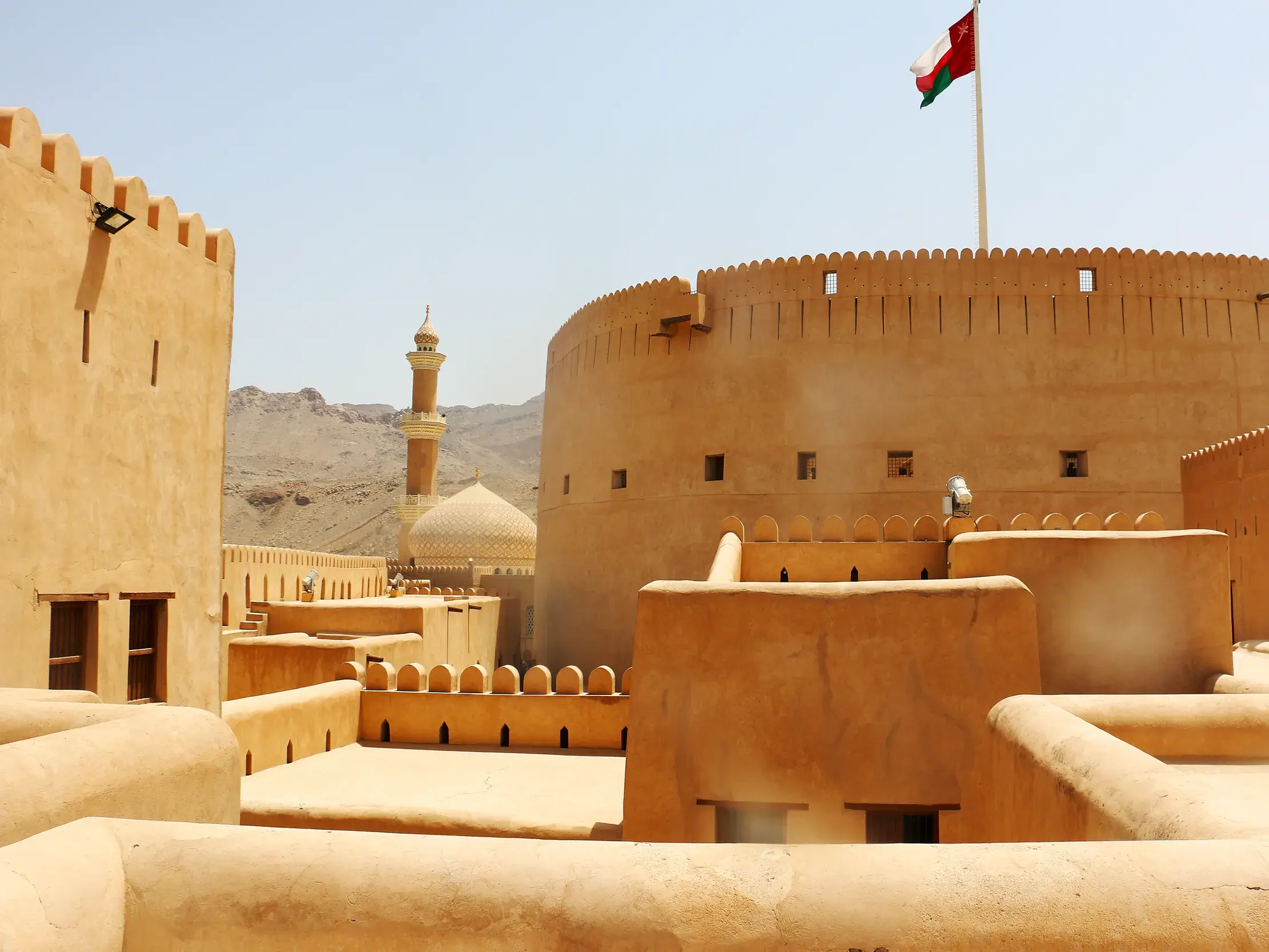 Imposanter Rundturm der sandfarbenen Festung von Nizwa im Oman, über dem die Nationalflagge vor dem strahlend blauen Himmel weht.