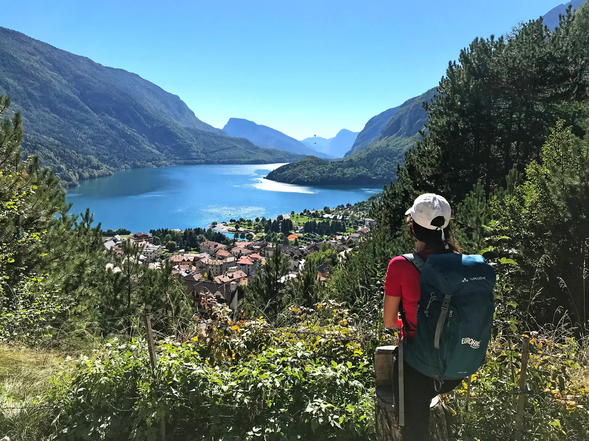 Eine Wanderin mit Rucksack macht eine Pause und blickt von einem Aussichtspunkt auf den türkisblauen Molvenosee im Trentino.
