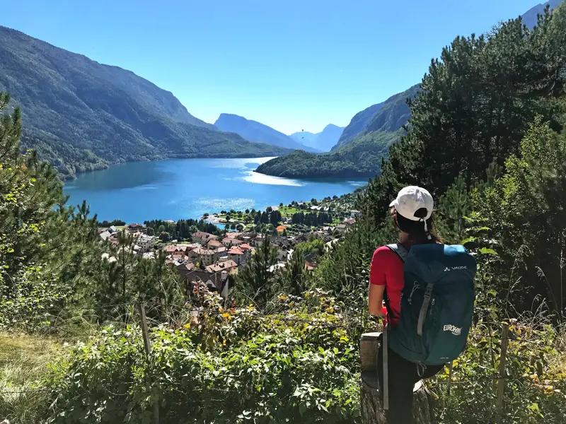Eine Wanderin mit Rucksack macht eine Pause und blickt von einem Aussichtspunkt auf den türkisblauen Molvenosee im Trentino.